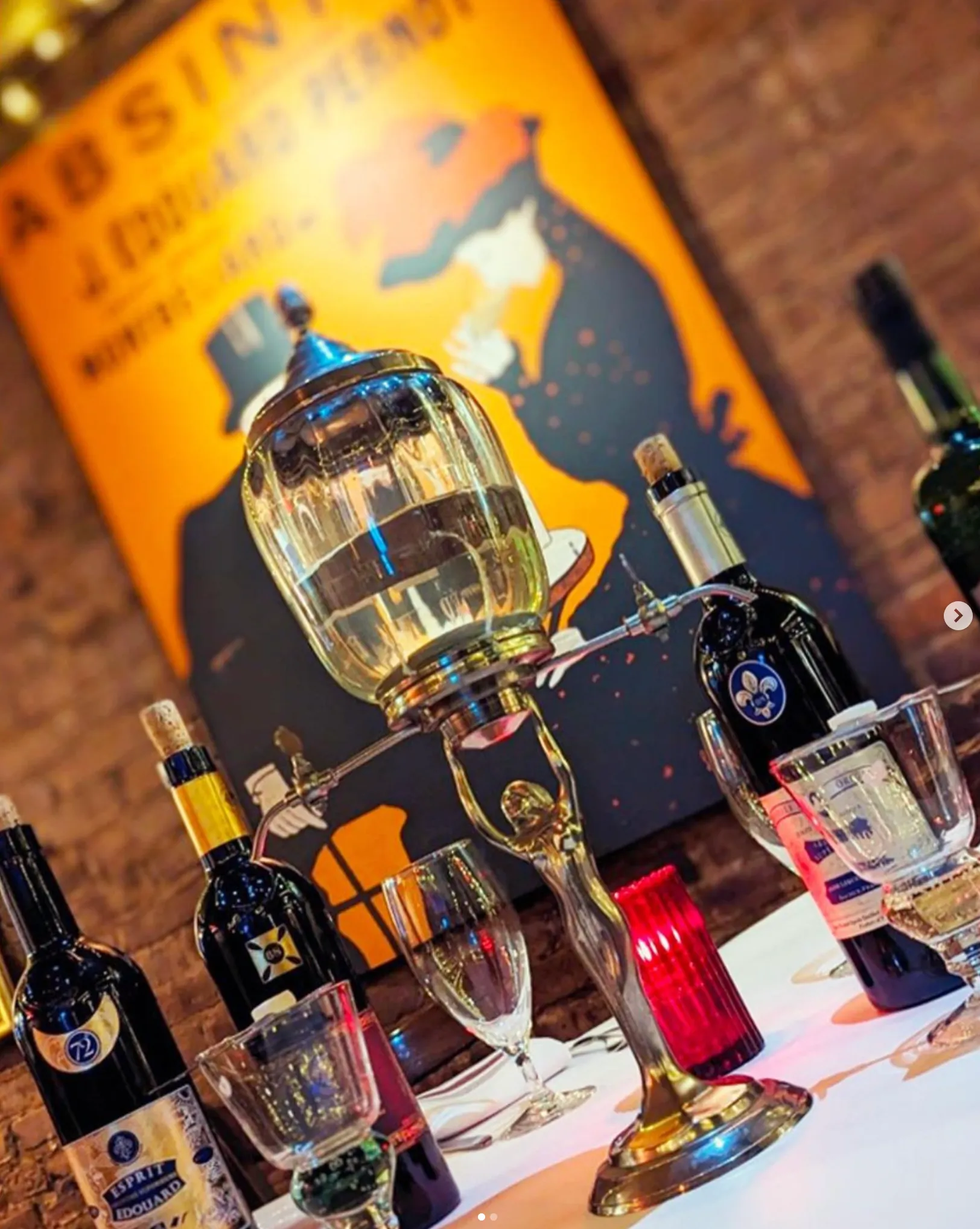 Absinthe fountain on a table surrounded by wine bottles and glasses with an Absinthe-themed poster in the background.