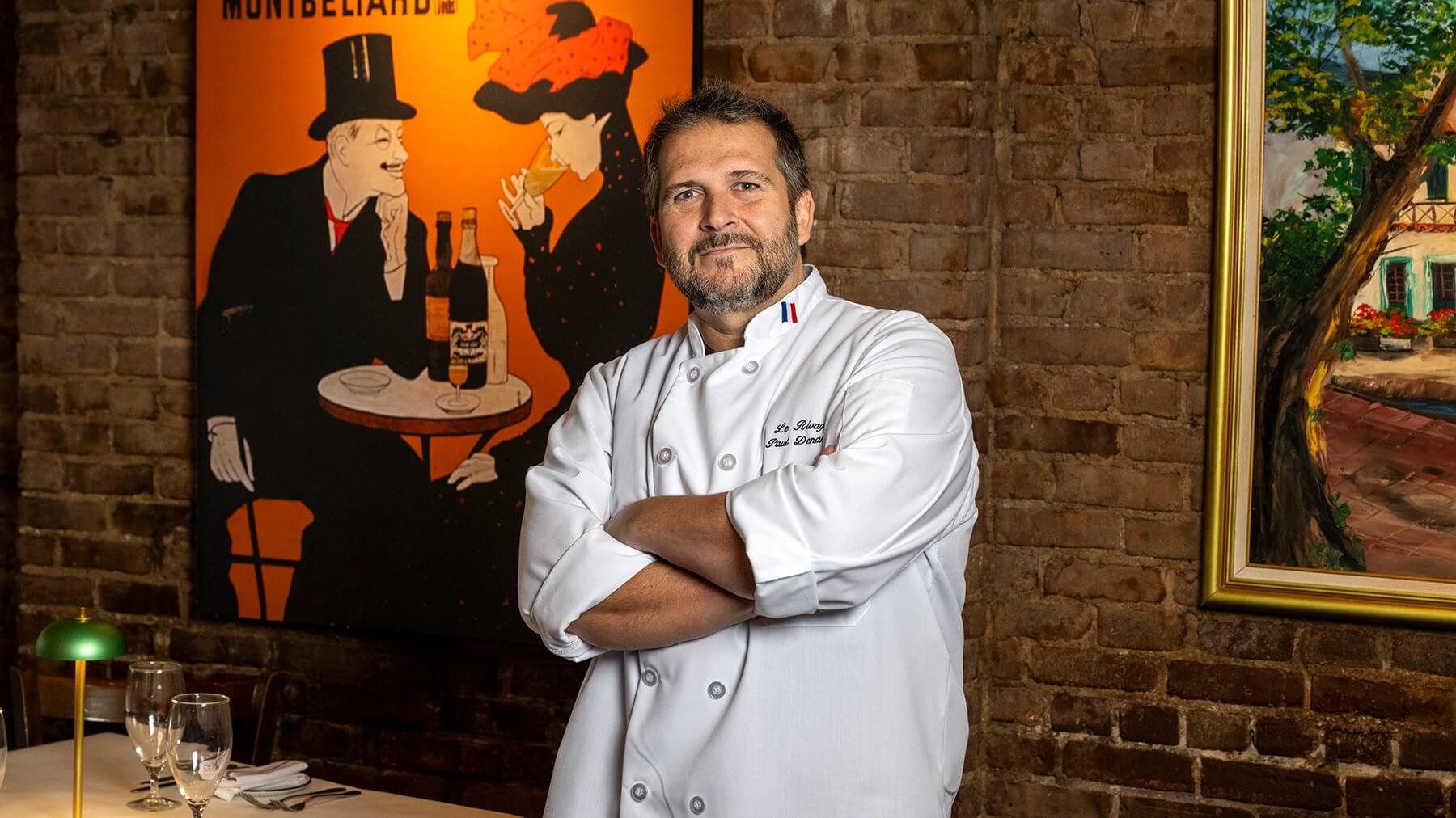 Chef in a white coat with folded arms standing against a brick wall with framed paintings.
