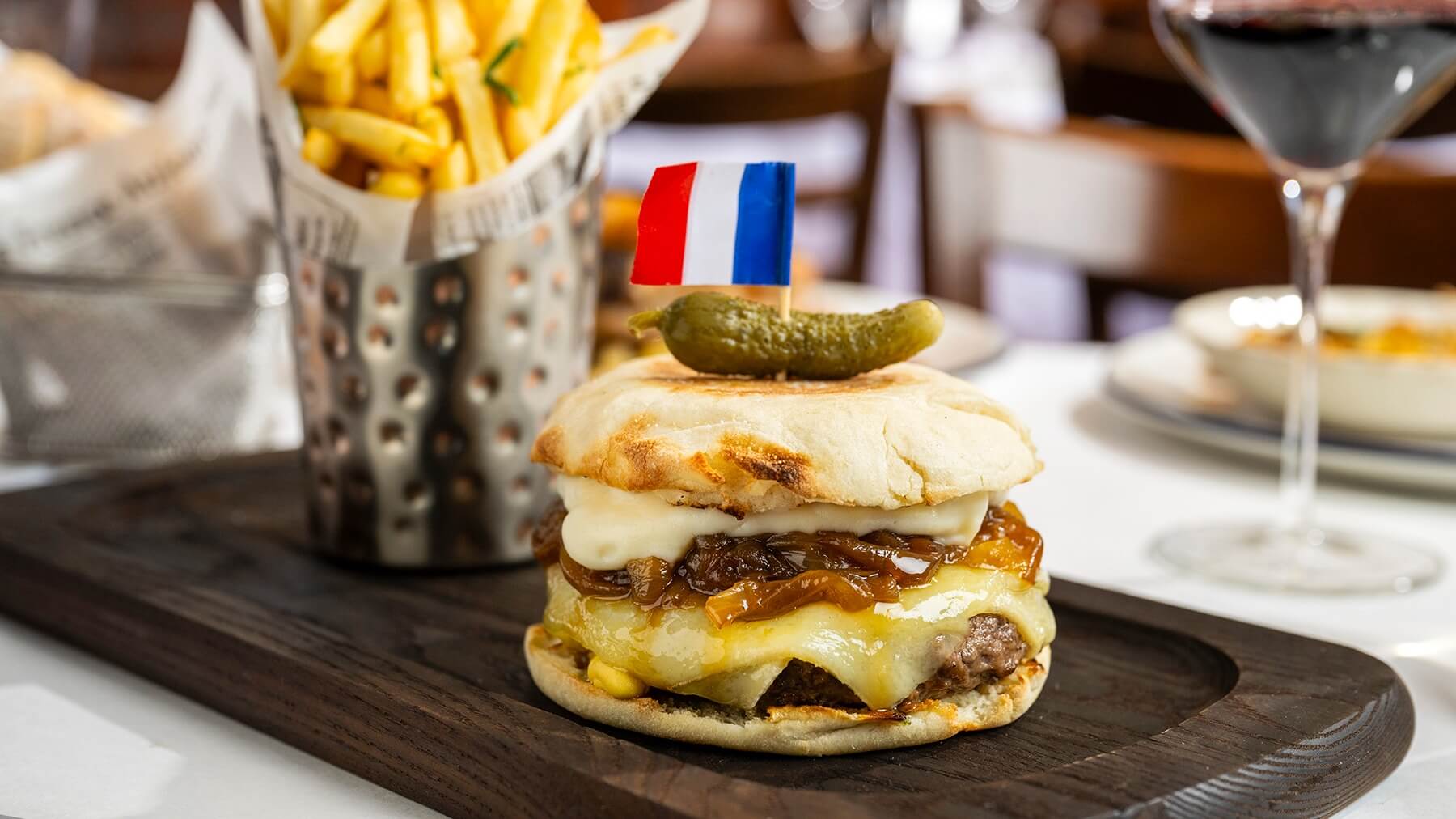 French onion soup burger topped with melted cheese, caramelized onions, melted cheese sauce, and a small pickle with a French flag toothpick, served on a wooden platter with a side of fries in the background.
