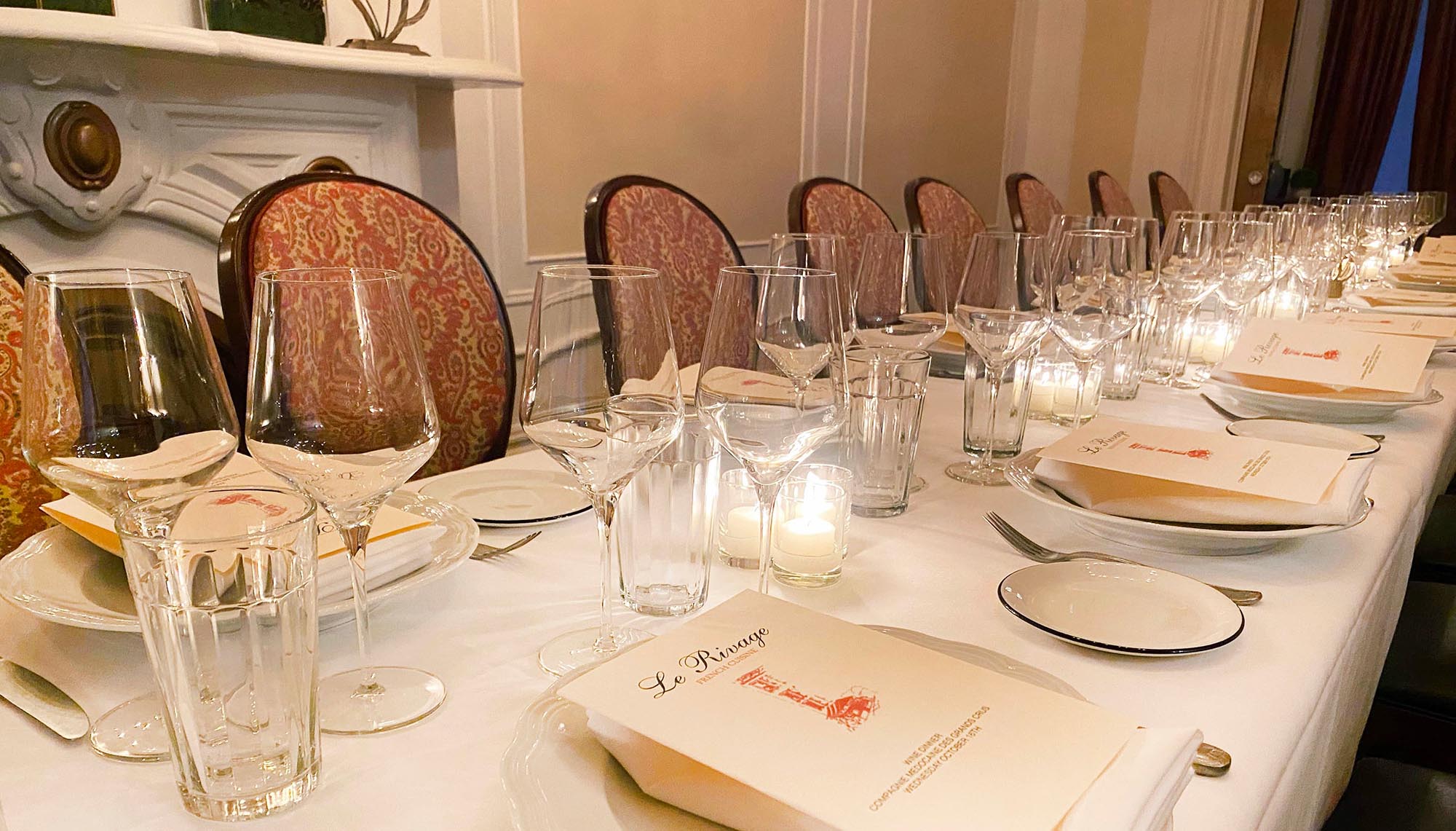 Long dining table set with wine glasses, water glasses, plates, and menus in an elegant room with patterned chairs and candles.