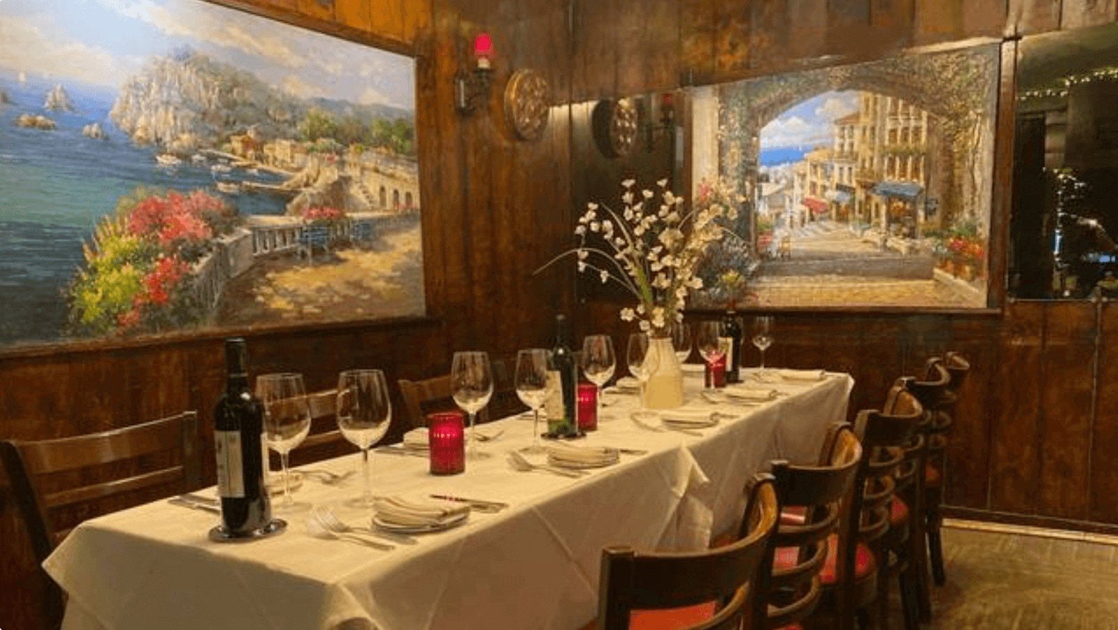 Elegant dining table in a wood-paneled room set with wine bottles, glasses, red candles, and a vase with white flowers, flanked by coastal-themed murals.