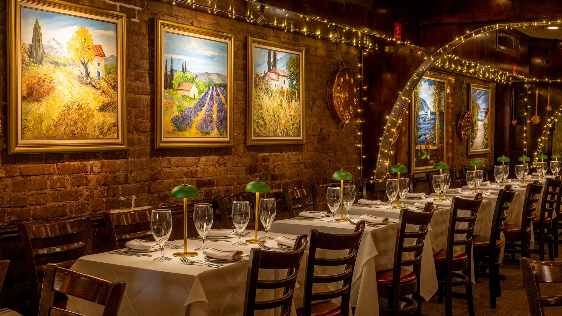 Elegant restaurant interior with long tables set with white tablecloths, glassware, green lamps, and framed landscape paintings on exposed brick walls decorated with string lights.